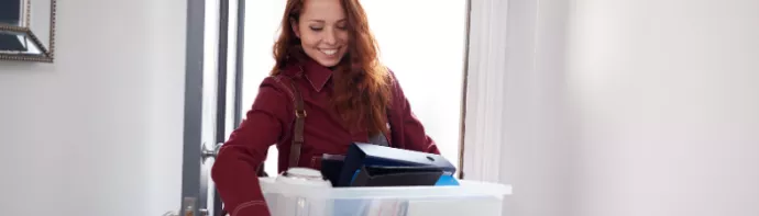 Smiling student enters door with a box of stuff in their arms.