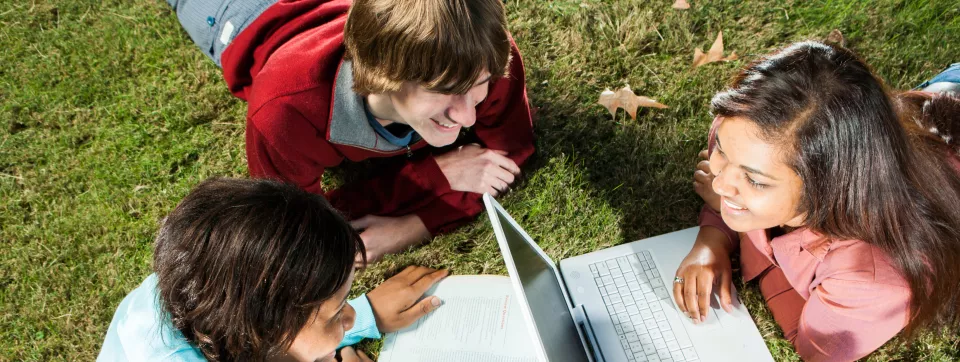 Three students sitting on the grass with laptops and notebooks.