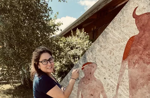 person holding a paintbrush next to a mural outside on a sunny summer day