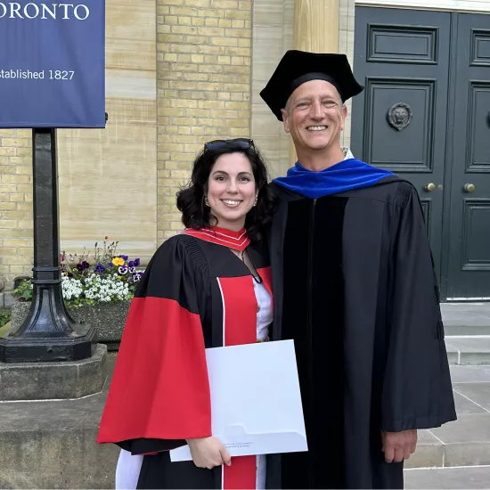 Dr. Paula Sanchez Nuñez de Villavicencio and Dr Jeremy Packer, in full academic robes, standing in front of Convocation Hall at the University of Toronto.