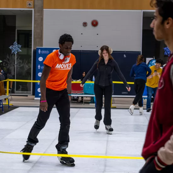Photo of a male individual in an orange shirt that says "UTM Moves" in white skating on a glice rink
