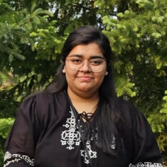 irtaqa arif smiling infront of shrubbery with a black, flowy long sleeve shirt with white stitched detailing