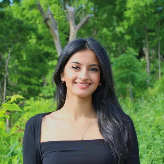 keerath bhachu, a brown woman, is wearing a black shirt, with long black hair draped over one shoulder, smiling in front of a bright green forest background