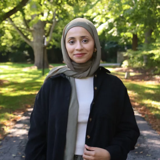 Areej smiling infront of a forest background, she is wearing a light brown hijab and black jacket