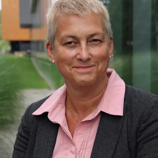 Angela Lange smiling in front of a building on the UTM campusm wearing a black blazer with a pink blouse underneath. She has short, grey hair. 