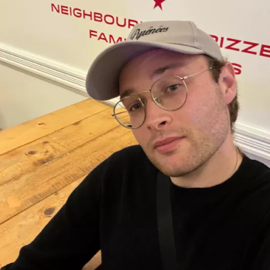 Dominik Jaworski smiling in a selfie style photo. He is wearing round glasses with a beige ball cap and plain black long sleeve shirt. He is sitting on a bench inside of a pizzeria. 