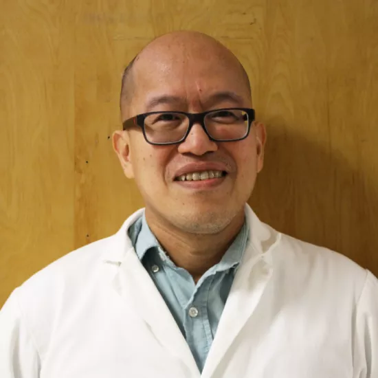 Windsor Chan headshot. He is wearing a white lab coat with a light blue collared button up underneath, as well as black, square glasses. He is standing in front of a light brown wooden door. 