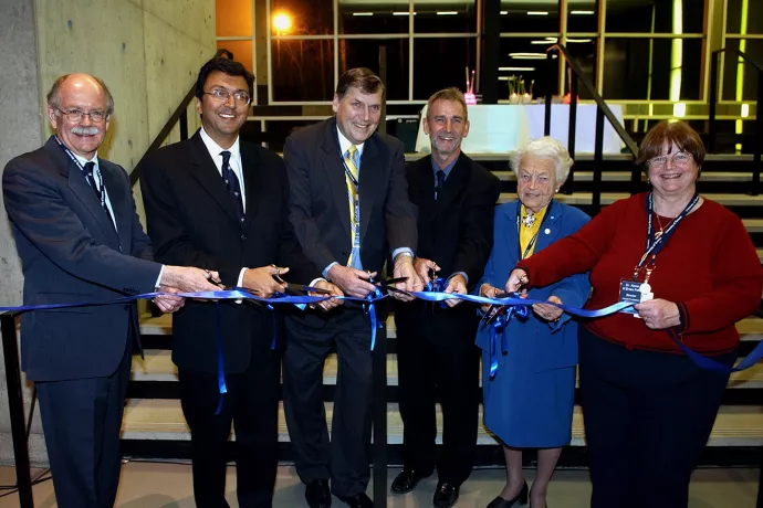 Four men in black suits standing in a line next to a woman in a blue dress suit and a woman in a red shirt, all holding a blue ribbon in front of them, each with a pair of scissors opened, ready to cut the ribbon.