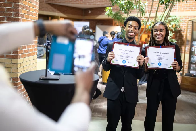 A man and woman in black suits stand together, each holding a certificate in front of them whilst smiling. In the foreground, out of focus, are arms and hands holding a cellphone taking a photo of the pair.