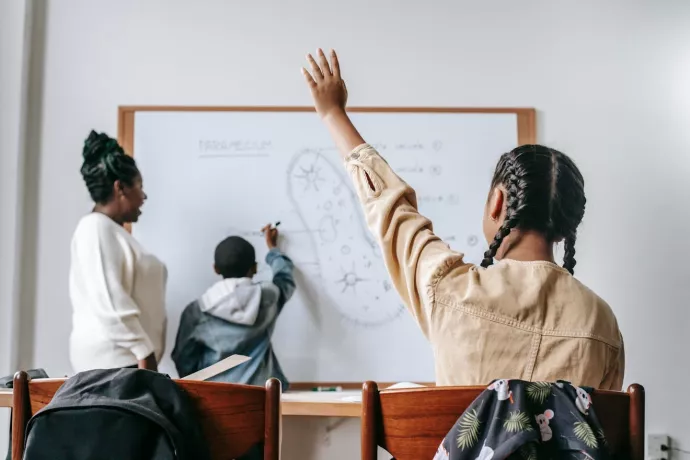 Child raises hand as another works on whiteboard with teacher looking on