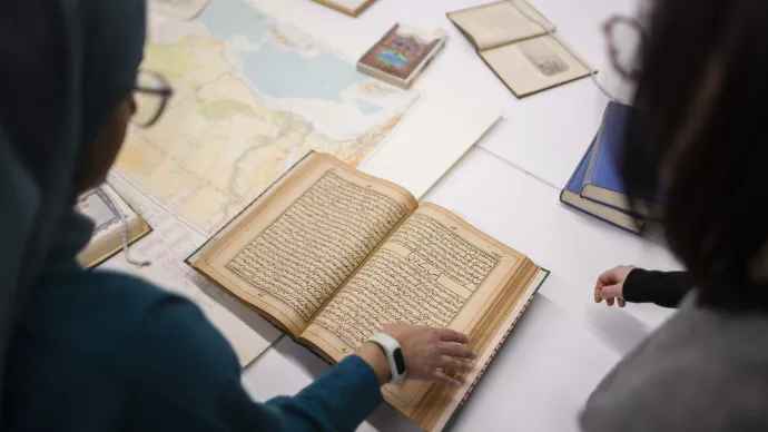 Two people look at an old book on a table next to a map of the world and other scattered old books