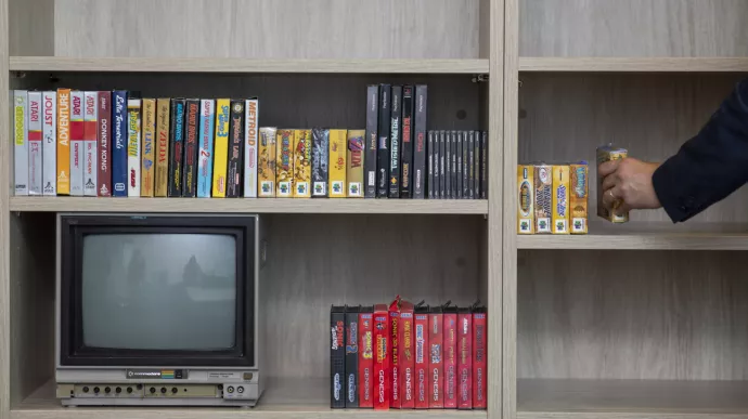 : A wooden shelving unit displays a collection of retro video games in their original boxes, including Atari, Nintendo 64, PlayStation, and Sega Genesis titles. A vintage Commodore monitor sits on the lower shelf. On the right side, a person’s hand is seen placing a boxed Nintendo 64 game on the shelf.