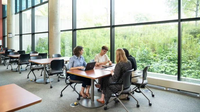 Four people sit around a table in a bright, modern study space with large windows overlooking lush greenery. They are engaged in discussion, using a laptop and notebooks. Other tables and chairs are visible in the background.