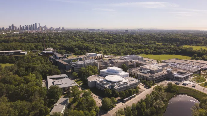  An aerial view of the University of Toronto Mississauga campus, surrounded by lush greenery, with the Mississauga skyline in the background. The modern academic buildings, parking lots, and nearby pond are visible under a clear summer sky.