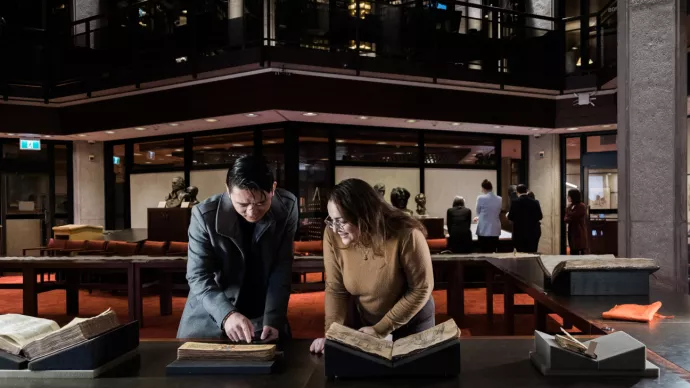 Two people examine an old manuscript in a dimly lit library. The person on the left, wearing a dark coat, points at the text, while the person on the right, wearing glasses and a brown sweater, smiles as they look at the book. Other visitors in the background view historical documents in a warmly lit, wood-panelled space with red carpeting and glass walls.