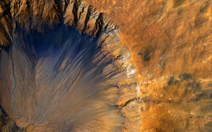 A high-resolution aerial image of a crater on Mars, displaying rugged, steep ridges with dark blue and orange hues. The terrain transitions from deep shadows inside the crater to a sunlit, rocky Martian surface.