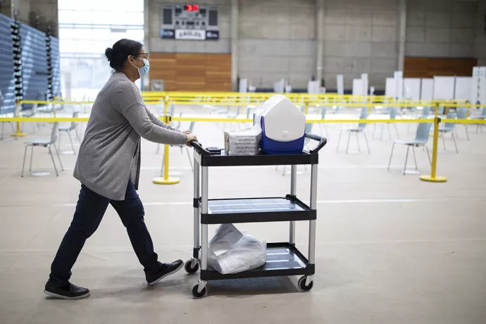 Woman pushes cart of medical supplied through RAWC gymnasium, which has rows of chairs physically distanced