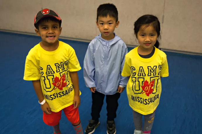 Three children standing in a gym, two wearing bright yellow t-shirts that read Camp U of T Mississauga