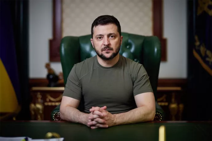 Volodymyr Zelenskyy wears an army-green shirt and sits at a desk in a leather-backed chair. The yellow-and-blue Ukrainian flag is visible in one corner.