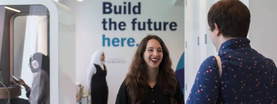 Two students talk and smile, in the background is a poster saying Build the future here.