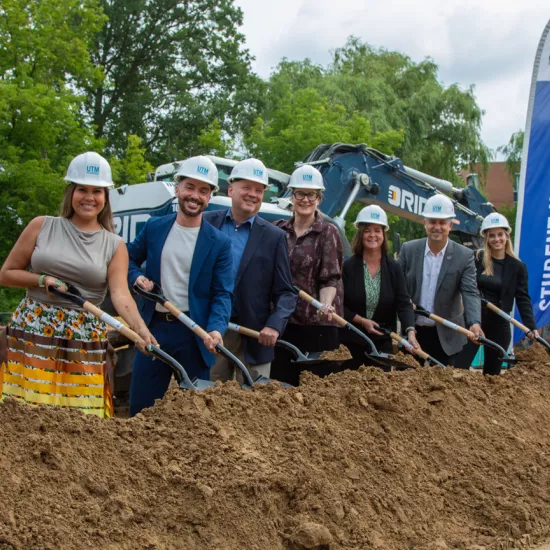 A group of people in hardhats holding shovels dig into dirt at groundbreaking ceremony for new residence. 