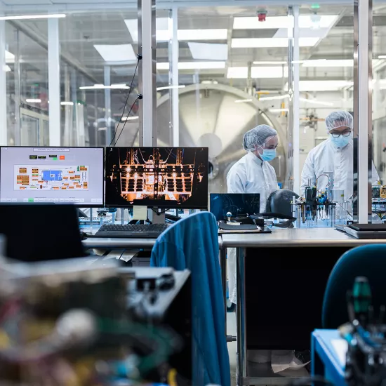 A lab with two computer monitors in the foreground in front of a glass wall. Behind the glass wall are two people dressed in white lab coats, hair nets and blue face masks with electronic equipment in front of them on a table.