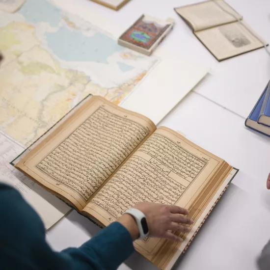 Two people look at an old book on a table next to a map of the world and other scattered old books