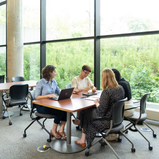 Four people sit around a table in a bright, modern study space with large windows overlooking lush greenery. They are engaged in discussion, using a laptop and notebooks. Other tables and chairs are visible in the background.