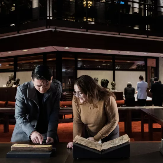 Two people examine an old manuscript in a dimly lit library. The person on the left, wearing a dark coat, points at the text, while the person on the right, wearing glasses and a brown sweater, smiles as they look at the book. Other visitors in the background view historical documents in a warmly lit, wood-panelled space with red carpeting and glass walls.