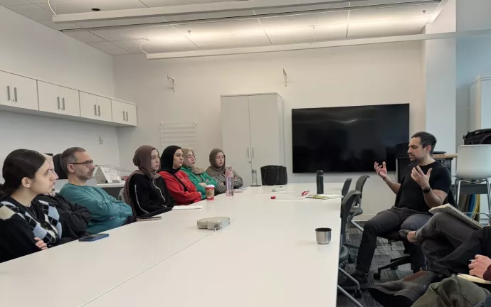 Professor Loubani presents in front of a crowd of students and professors around a boardroom table in an academic lab.