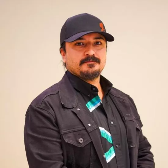 A man with a beard and mustache, wearing a baseball cap, black button-up flannel with a patterned shirt underneath.